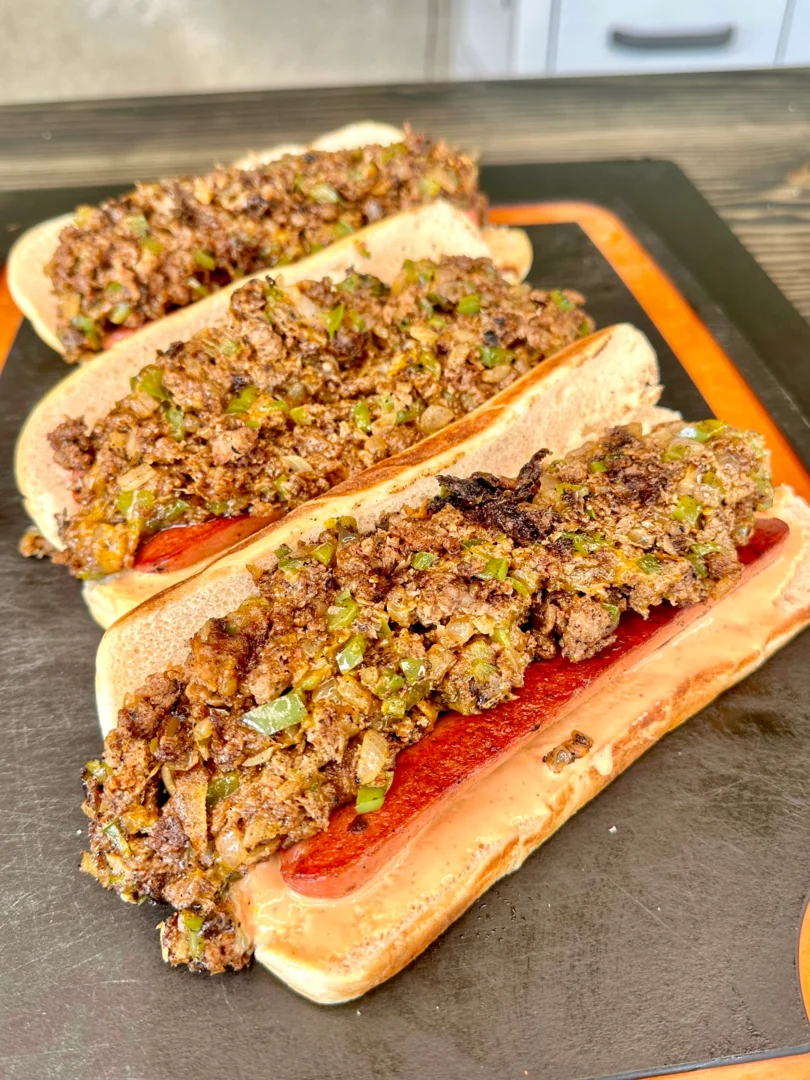 3 cheesesteak loaded hot dogs on an epicurian cutting board
