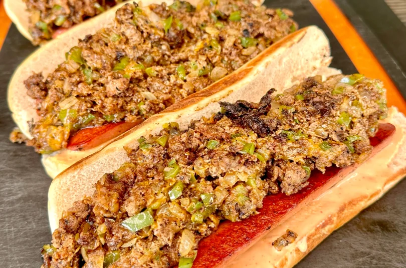 3 cheesesteak loaded hot dogs on an epicurian cutting board