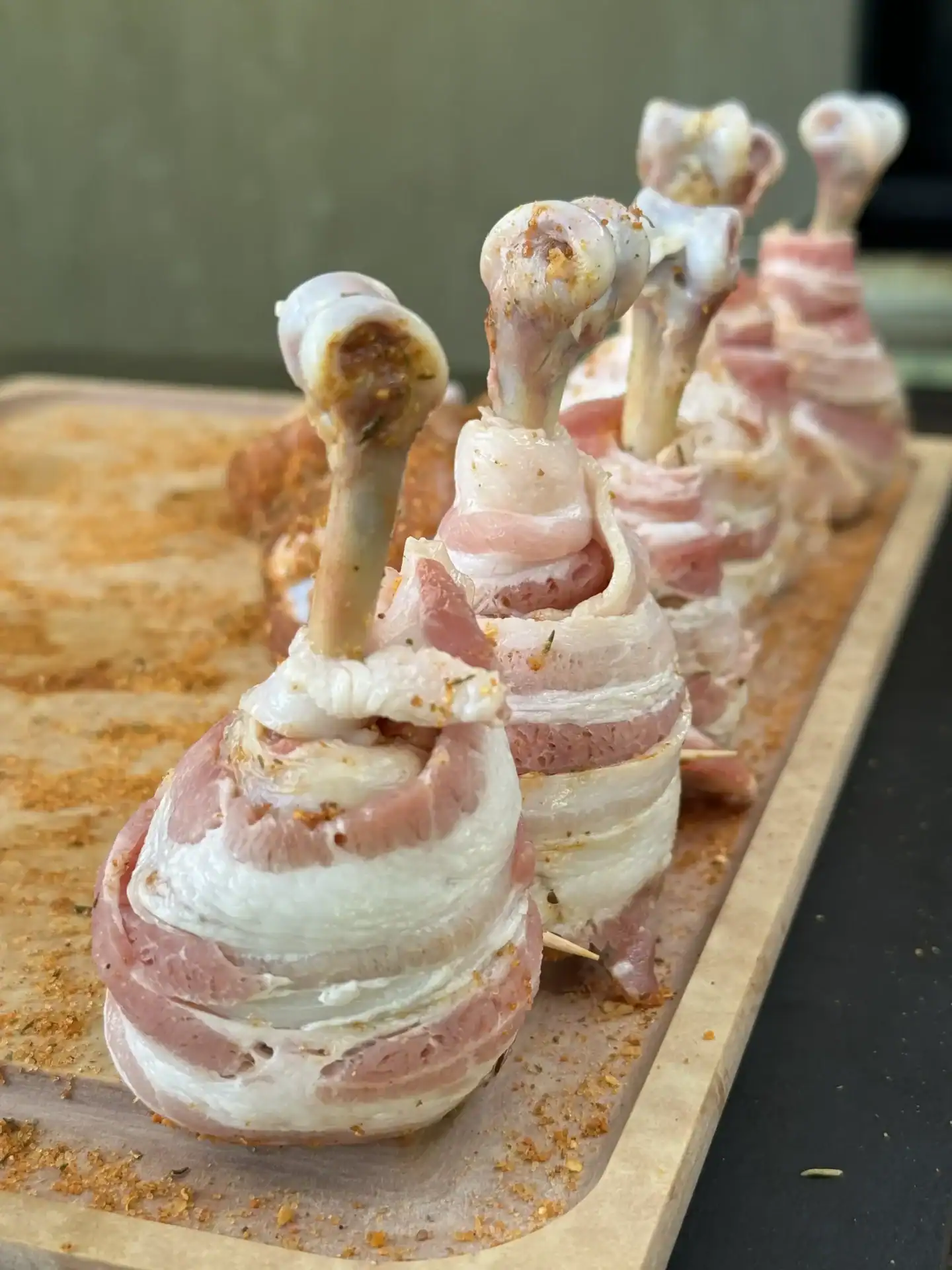 Seasoned chicken lollipops wrapped in regular-cut bacon and standing upright on a baking sheet before smoking.