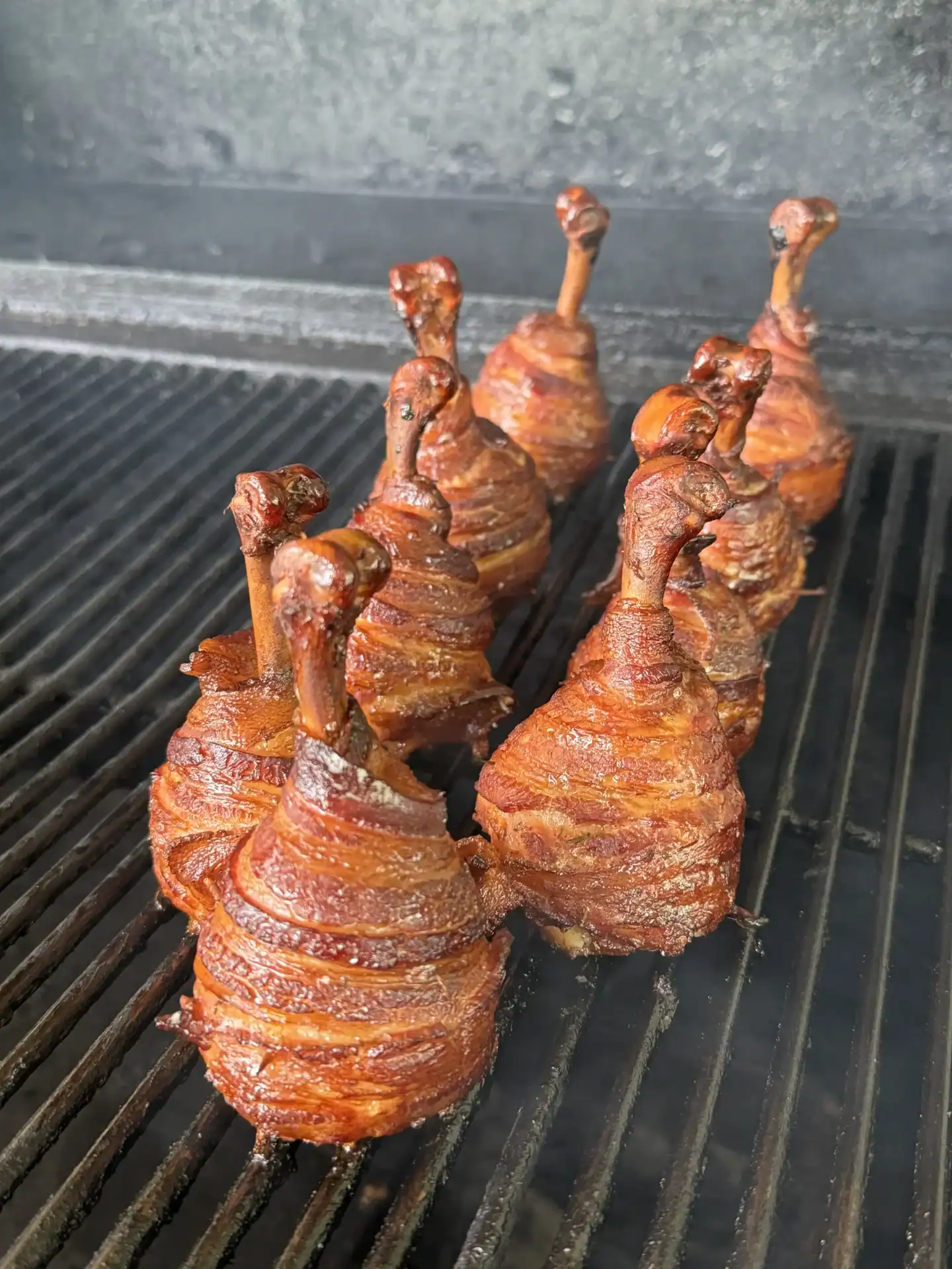 Bacon-wrapped chicken lollipops smoking upright on grill grates with reddish-brown bacon beginning to render.