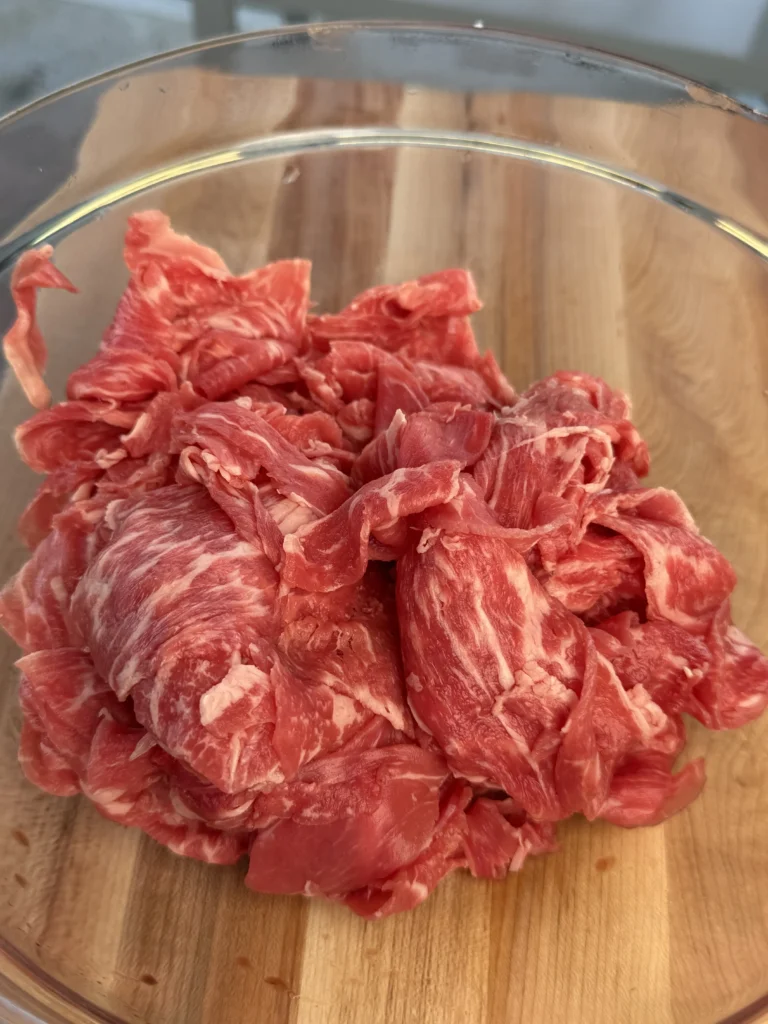 Close-up of raw shaved ribeye steak in a bowl board, ready for seasoning.
