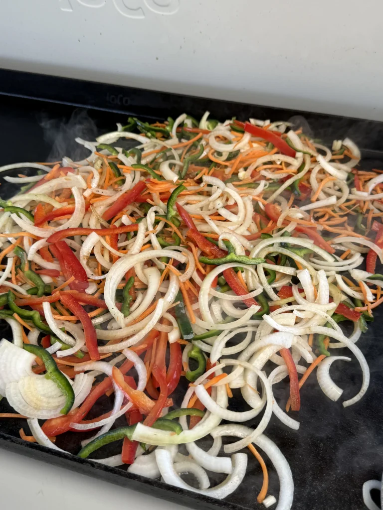 Peppers and onions sizzling on the griddle, showing light charring.
