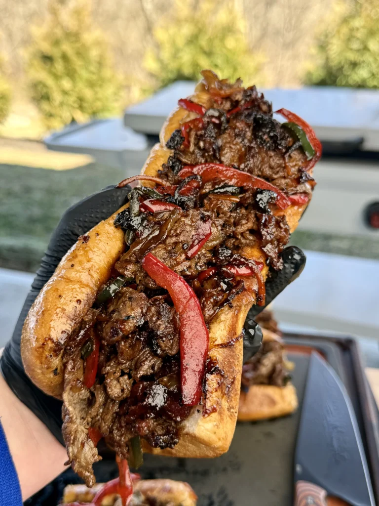 A person holding a griddled cheesesteak, ready to take a bite, with melted cheese stretching from the roll.