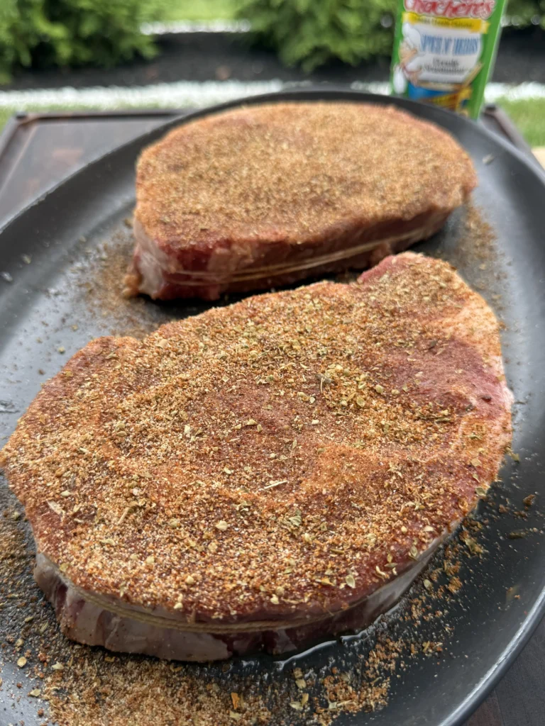 Two ribeye steaks heavily coated with brown and golden Cajun blackening spice rub in black skillet, with Tony's seasoning visible in background