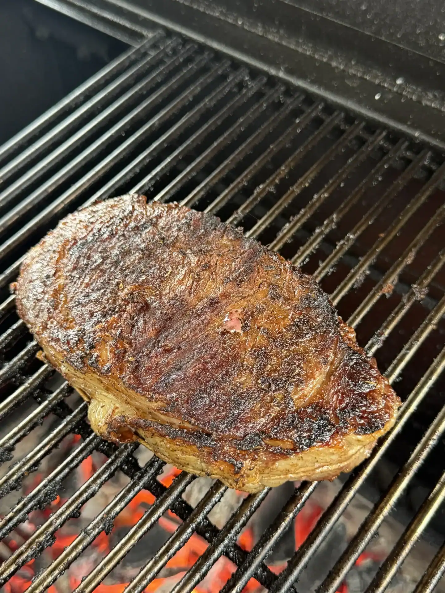 Single ribeye steak with dark blackened crust cooking on grill grates with orange flames visible, demonstrating perfect spice caramelization