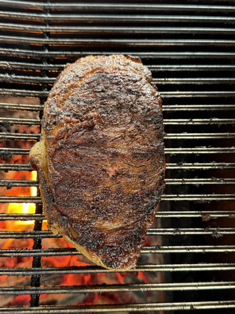 Different view of perfectly blackened ribeye steak on grill with dark caramelized spice coating and visible grill marks