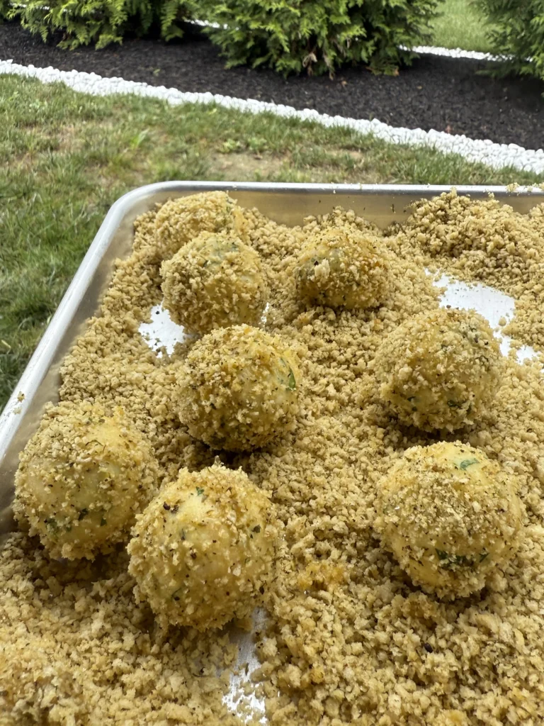 Multiple potato croquettes completely coated in golden breadcrumbs in aluminum tray, ready for deep frying with excess crumbs visible