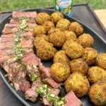 Complete steakhouse dinner showing sliced ribeye with herb garnish alongside golden crispy croquettes on black plate with Tony's seasoning