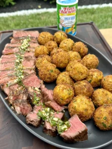 Complete steakhouse dinner showing sliced ribeye with herb garnish alongside golden crispy croquettes on black plate with Tony's seasoning