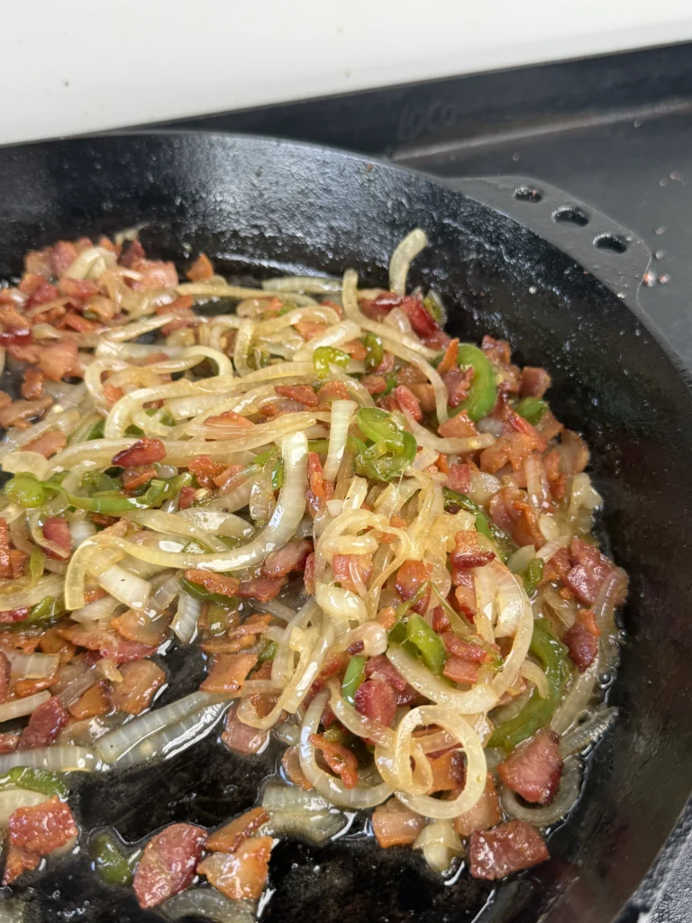 Crispy bacon pieces mixed with caramelized onions and jalapeños in black skillet, showing combination stage of bacon jam preparation