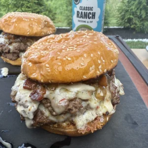 Several completed jalapeño bacon jam smash burgers arranged on slate board with ranch dressing bottle prominently displayed in background