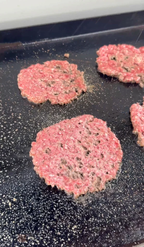 Four smashed raw beef patties spread thin on black cooking surface, showing ideal thickness and irregular edges for smash burgers