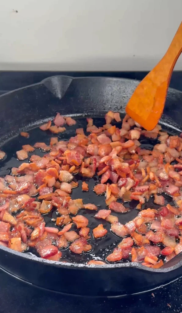 Chopped bacon pieces cooking and rendering fat in black skillet, showing preparation of bacon component for the jam