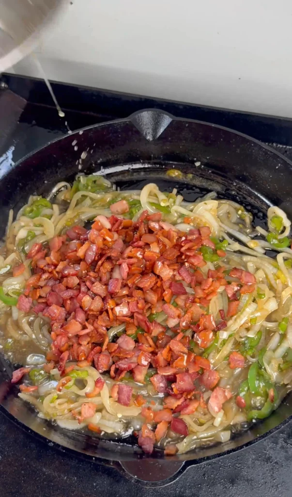 Cooked bacon being combined with sautéed onions and jalapeños in skillet, demonstrating final assembly stage of homemade bacon jam