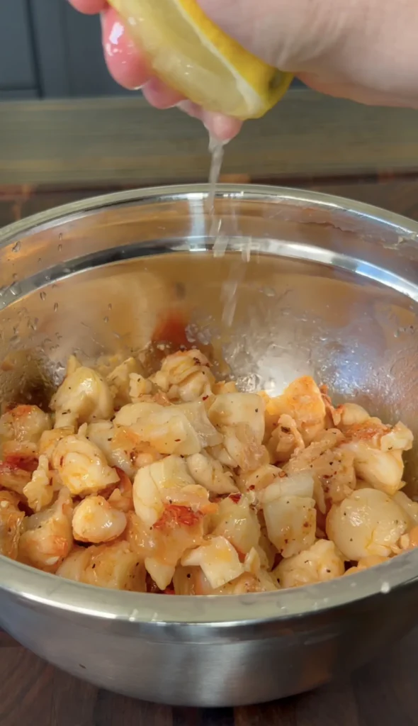Fresh lobster meat seasoned with spices in a stainless steel mixing bowl, being drizzled with sauce
