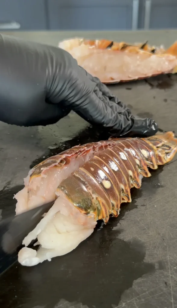 Chef in black gloves preparing fresh lobster tail with knife on stainless steel counter for hot honey butter lobster rolls