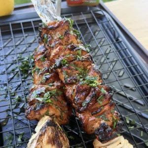Grilled marinated chicken skewers with herbs on a black grill rack alongside charred corn and onions