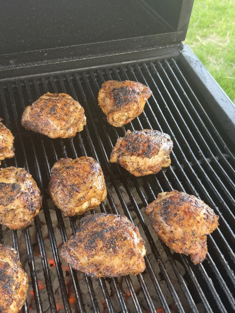 Seasoned honey chili crisp chicken thighs cooking on a black grill grate with charred golden-brown skin