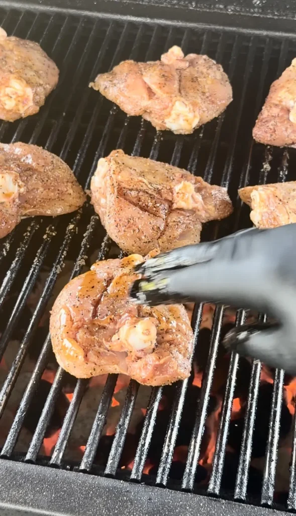 Honey chili crisp chicken thighs cooking on grill grates with tongs flipping one piece, showing golden-brown seasoned skin