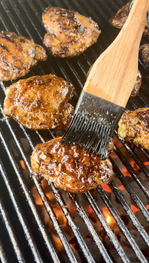 Honey chili crisp chicken thighs grilling on grates with wooden basting brush applying glaze over flames