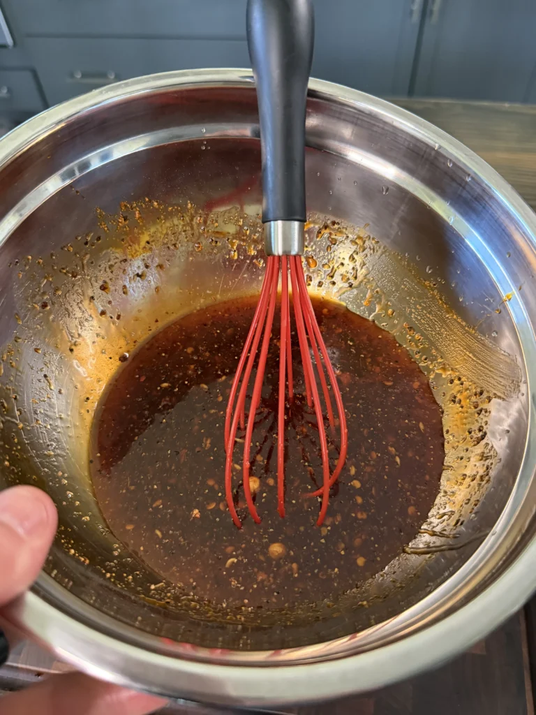 Dark honey chili crisp marinade being whisked in stainless steel bowl with red silicone whisk