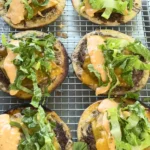 Six Oklahoma onion smash burger tacos with melted cheese, lettuce and sauce cooling on wire rack