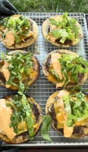 Six Oklahoma onion smash burger tacos with melted cheese, lettuce and sauce cooling on wire rack