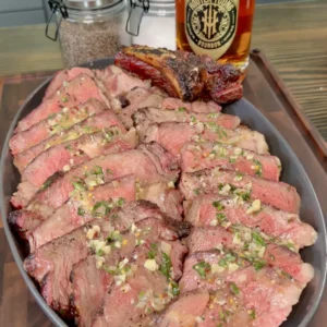 Sliced grilled steak topped with herb butter on gray platter with whiskey bottle in background