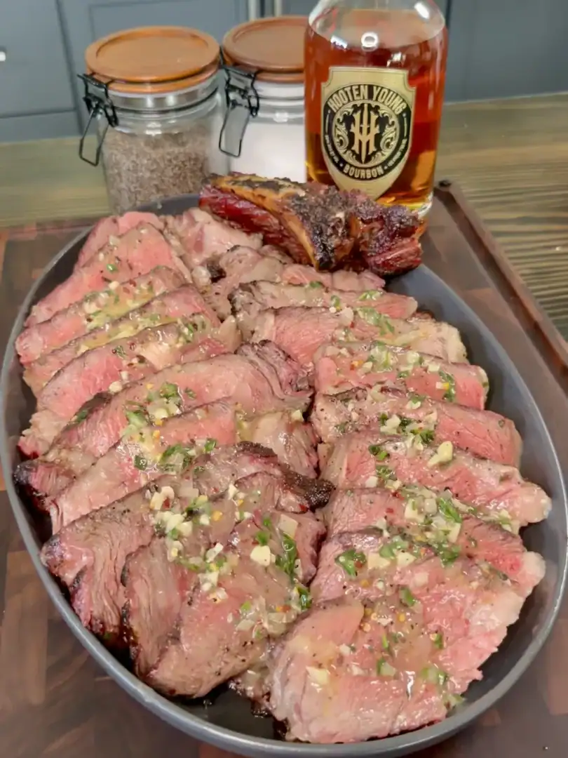 Sliced grilled steak topped with herb butter on gray platter with whiskey bottle in background