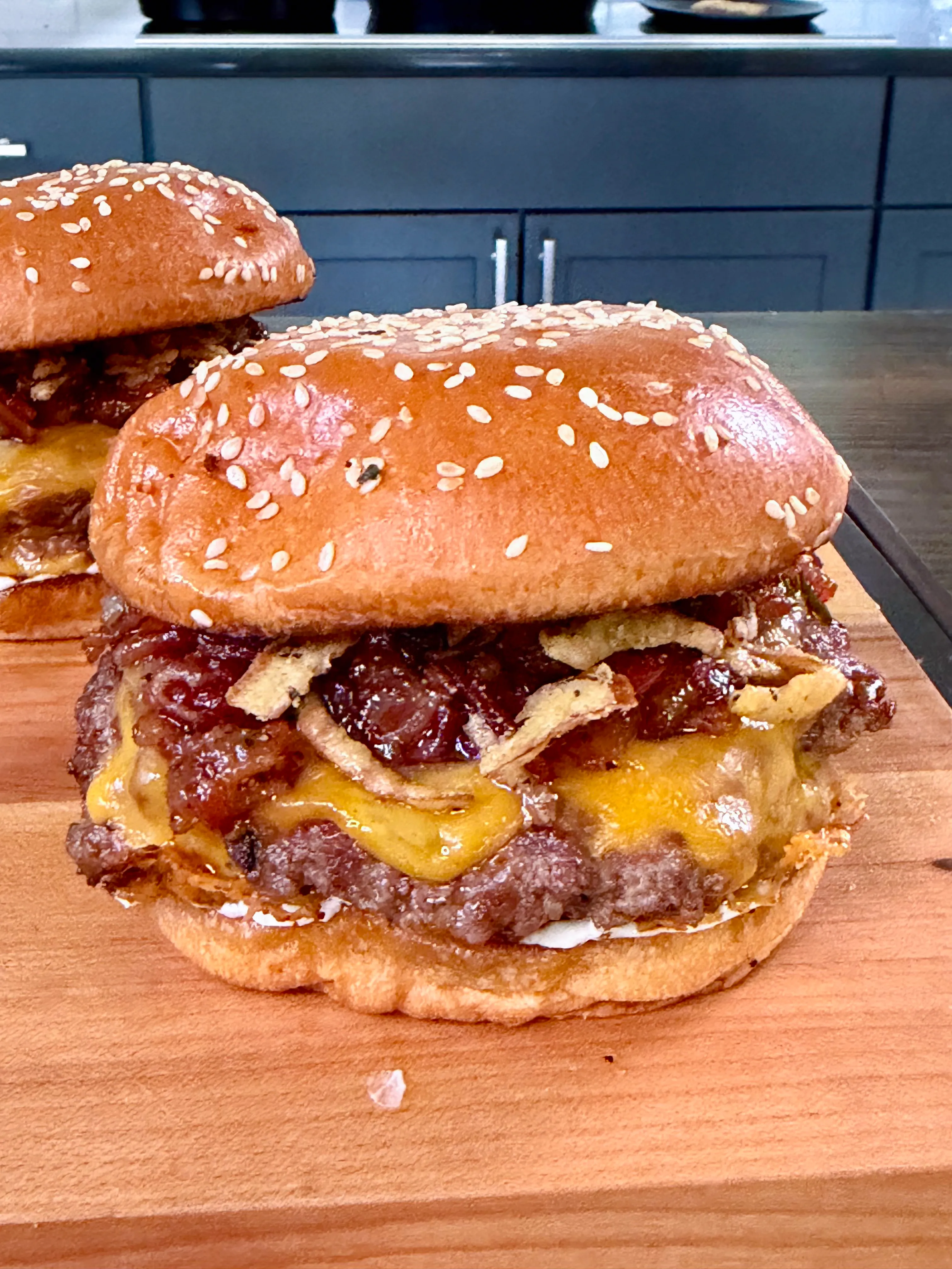 Two bacon jam smash burgers with melted cheese on sesame seed buns on a wooden cutting board