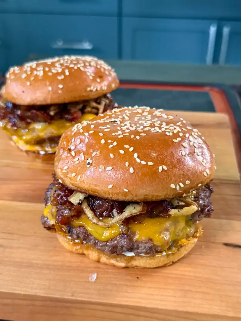 Two bacon jam smash burgers with melted cheese on sesame seed buns, served on a wooden cutting board