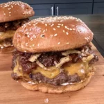 Two bacon jam smash burgers with melted cheese and caramelized onions on sesame seed buns served on wooden cutting board