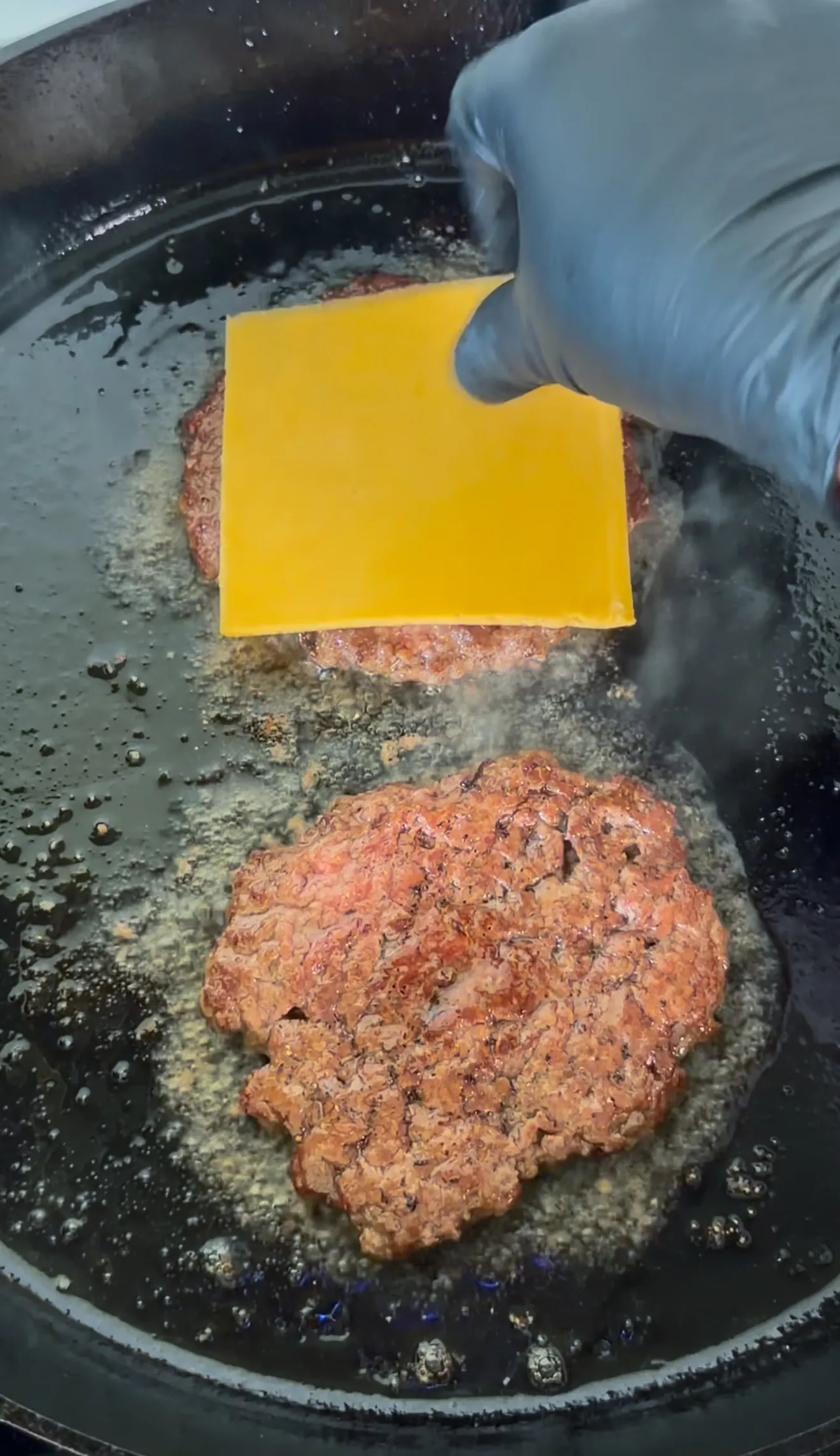 Two bacon jam smash burgers cooking in a cast iron pan with melted cheese being placed on top