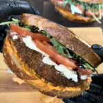 Crispy chicken cutlet sandwich on grilled sourdough bread with tomatoes, greens, and cheese held in black gloved hands