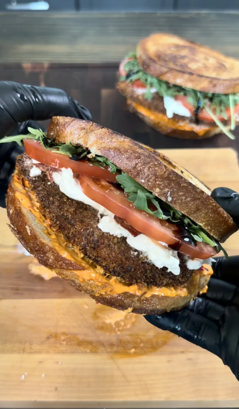 Crispy chicken cutlet sandwich on grilled sourdough bread with tomatoes, greens, and cheese held in black gloved hands