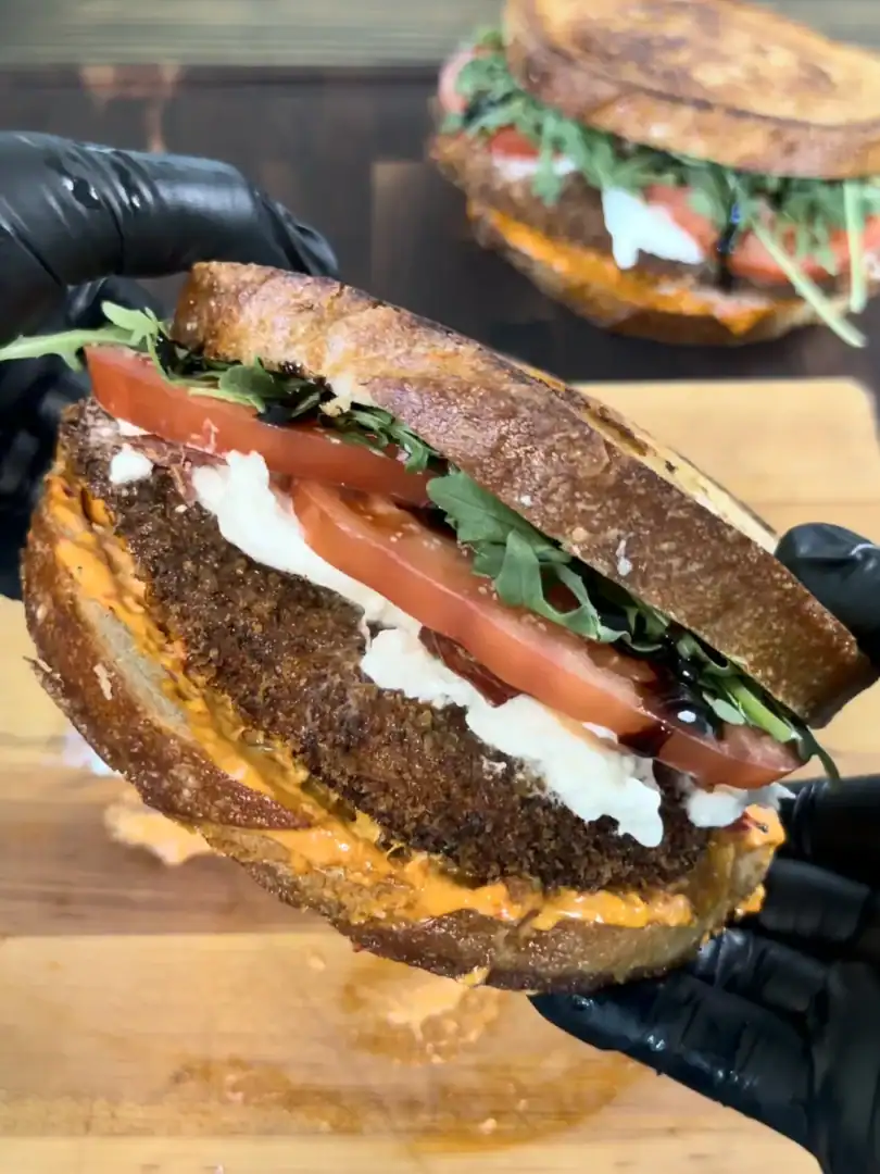 Crispy chicken cutlet sandwich on grilled sourdough bread with tomatoes, greens, and cheese held in black gloved hands