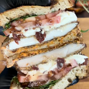Halved crispy chicken cutlet sandwich with melted cheese, bacon, and greens being grilled on black grill pan