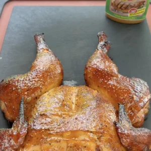 Golden brown spatchcock smoked chicken with crispy skin on a dark cutting board with seasoning jar