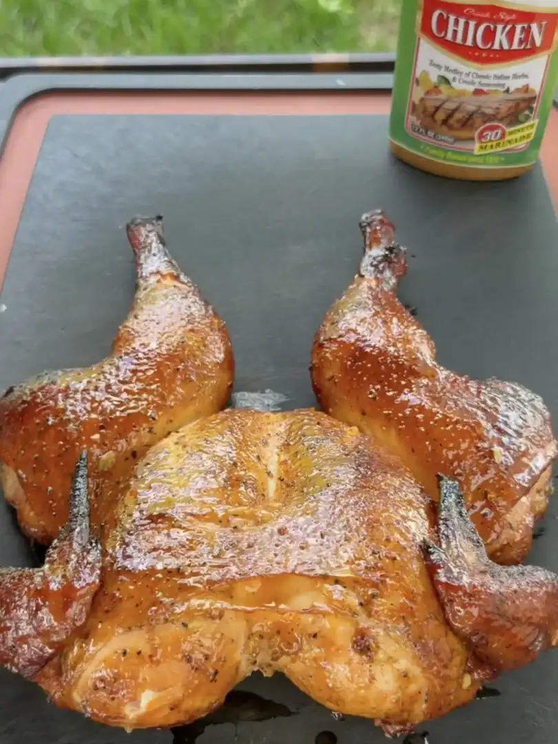 Golden brown spatchcock smoked chicken with crispy skin on a dark cutting board with seasoning jar