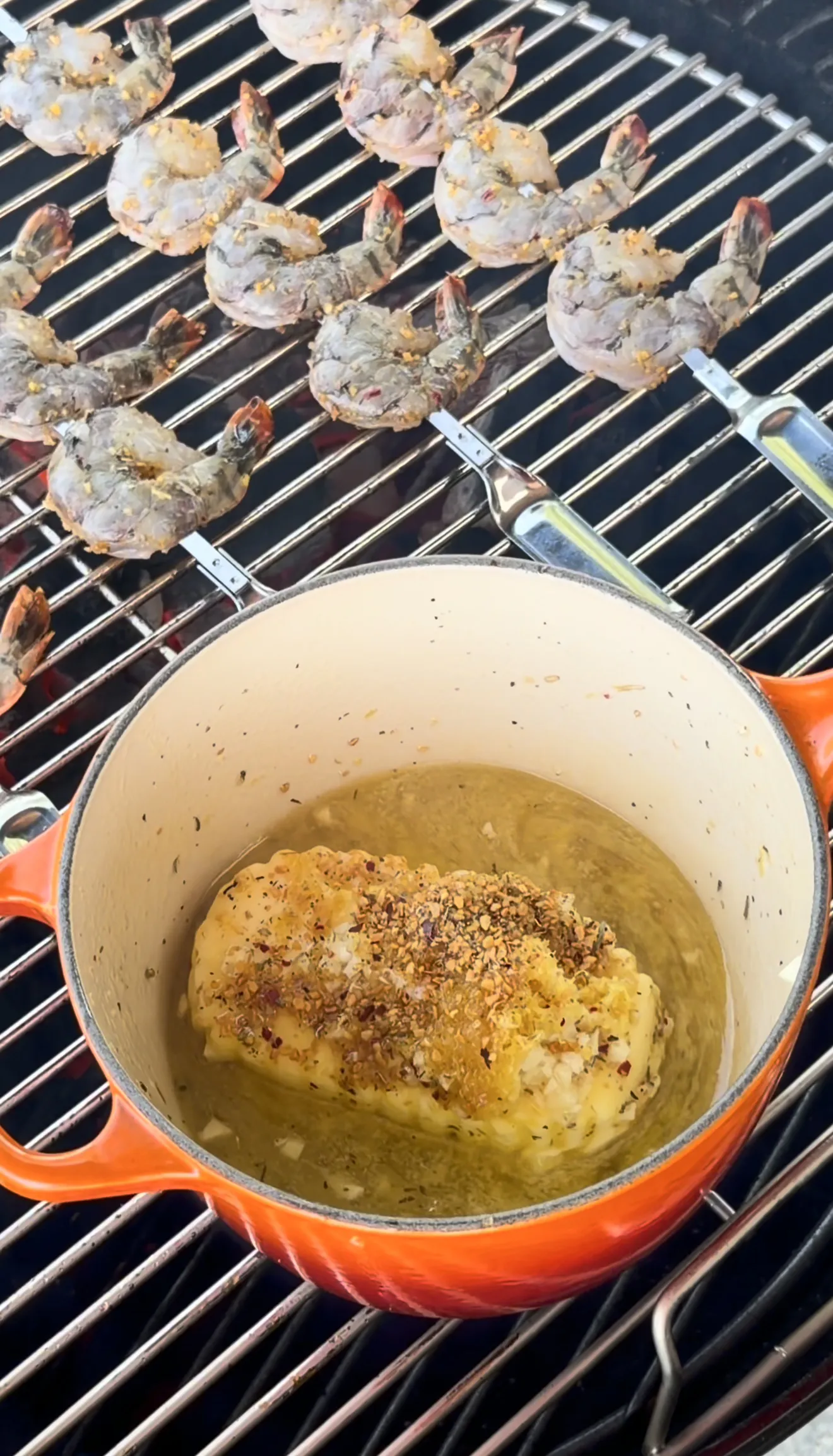 Grilled shrimp skewers on grill grates with roasted garlic herb butter in orange cast iron pot