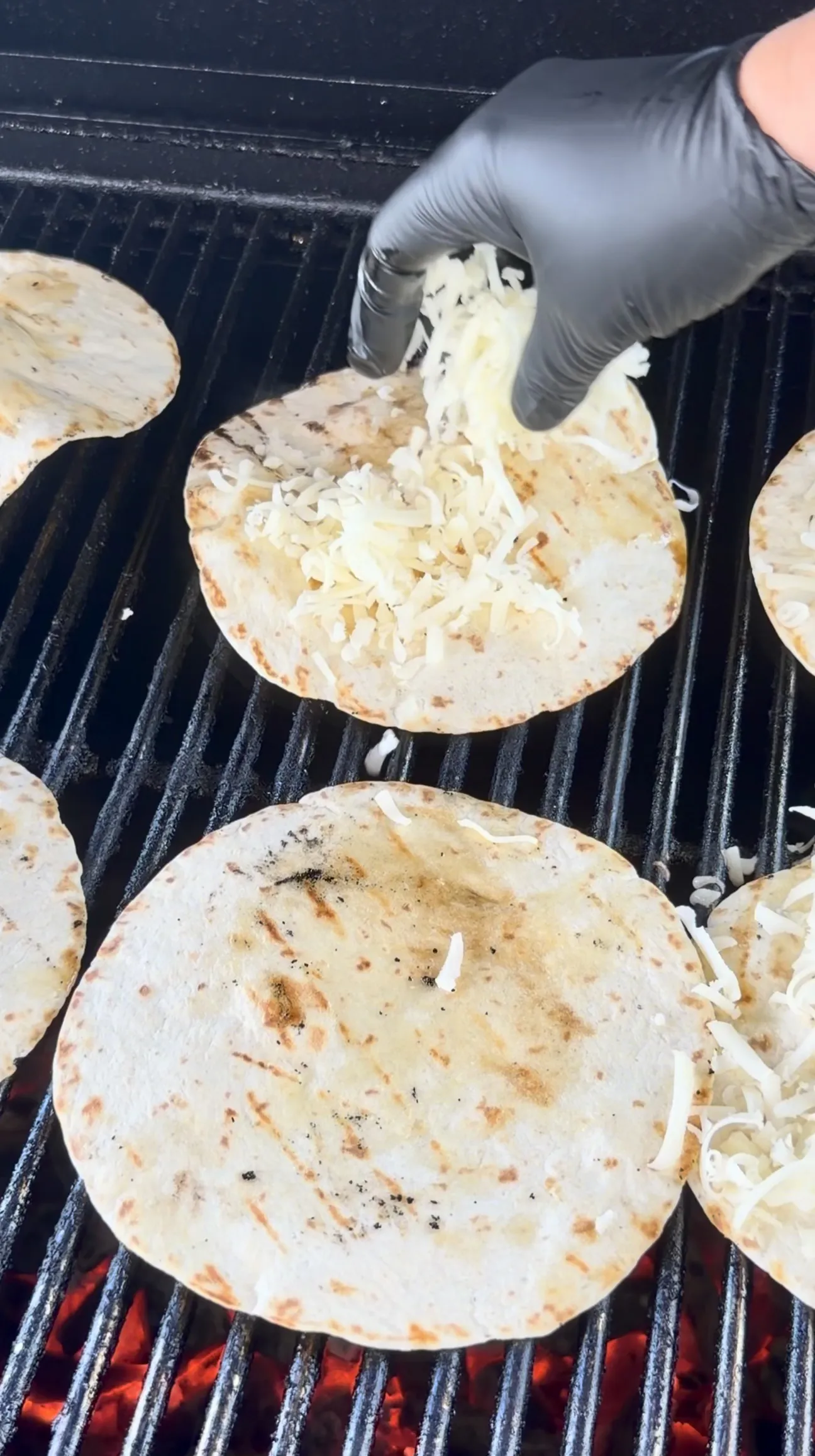Flour tortillas on grill grates with melted cheese being sprinkled on top by a gloved hand
