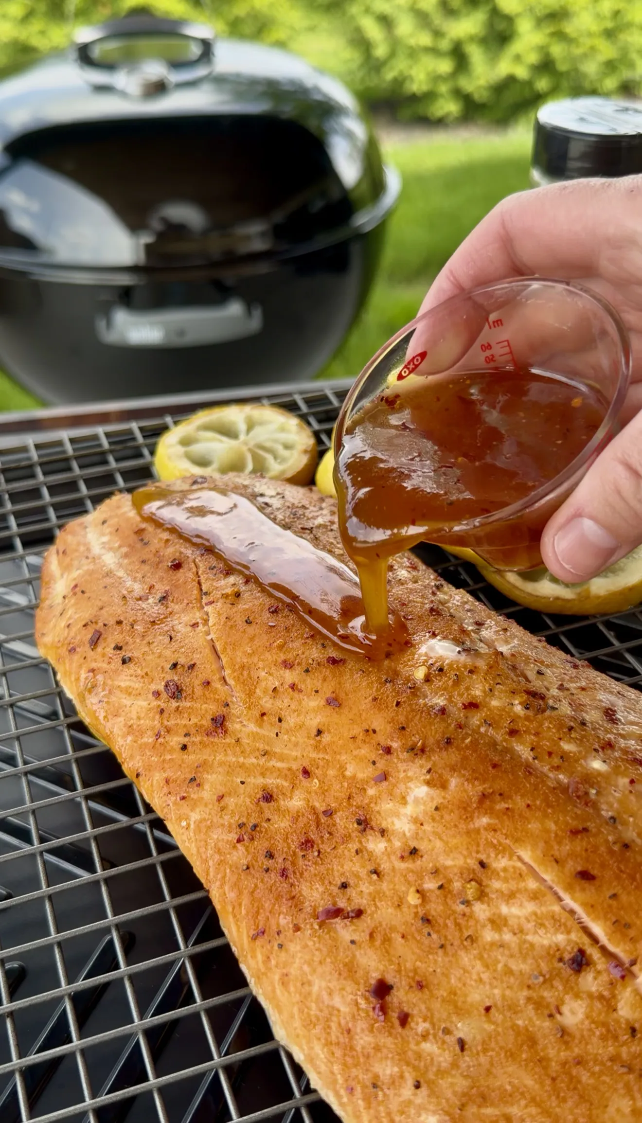 Hot honey being poured over seasoned smoked salmon fillet on grill grates with lemon slices