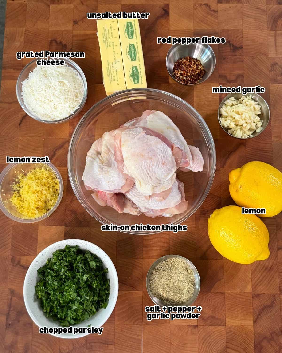 raw chicken thighs in a bowl, unsalted butter, lemon zest, lemons and other ingredients for grilled garlic Parmesan chicken thighs placed on a wooden chopping board.