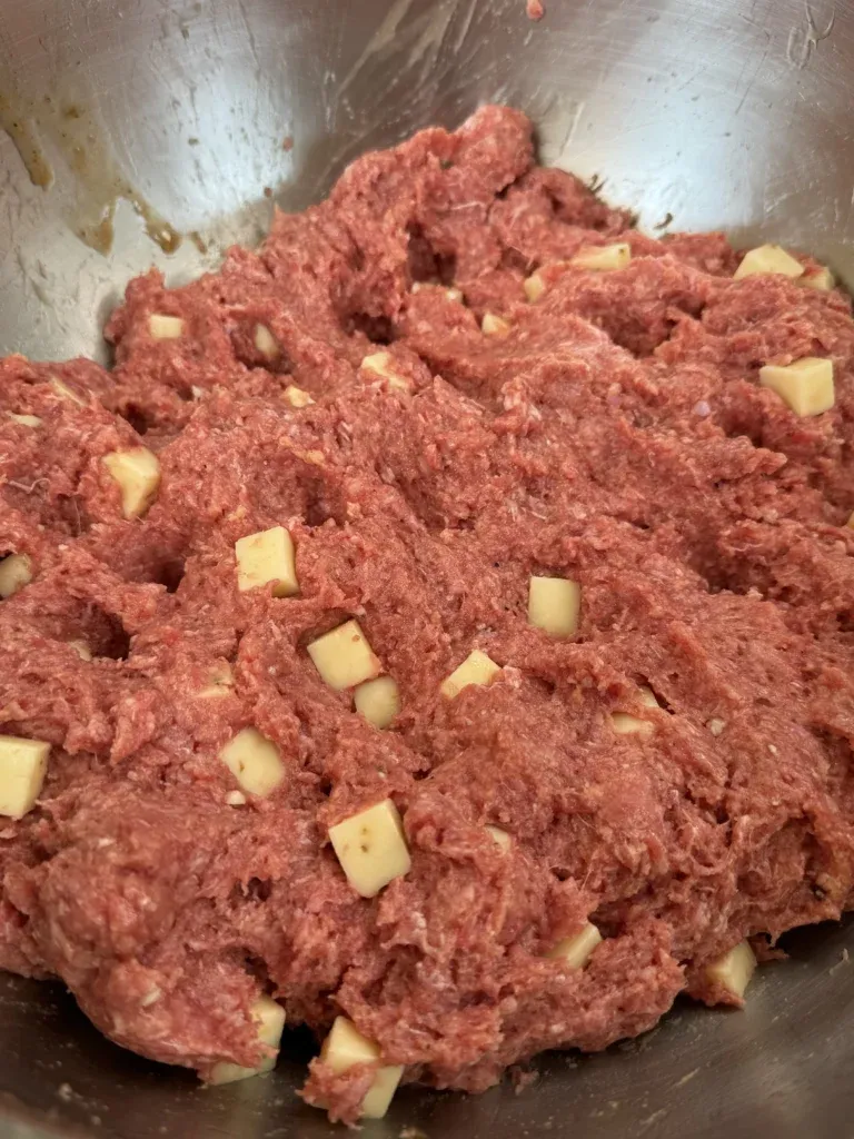 Raw ground beef mixed with small yellow cheese cubes in a stainless steel mixing bowl for burger preparation