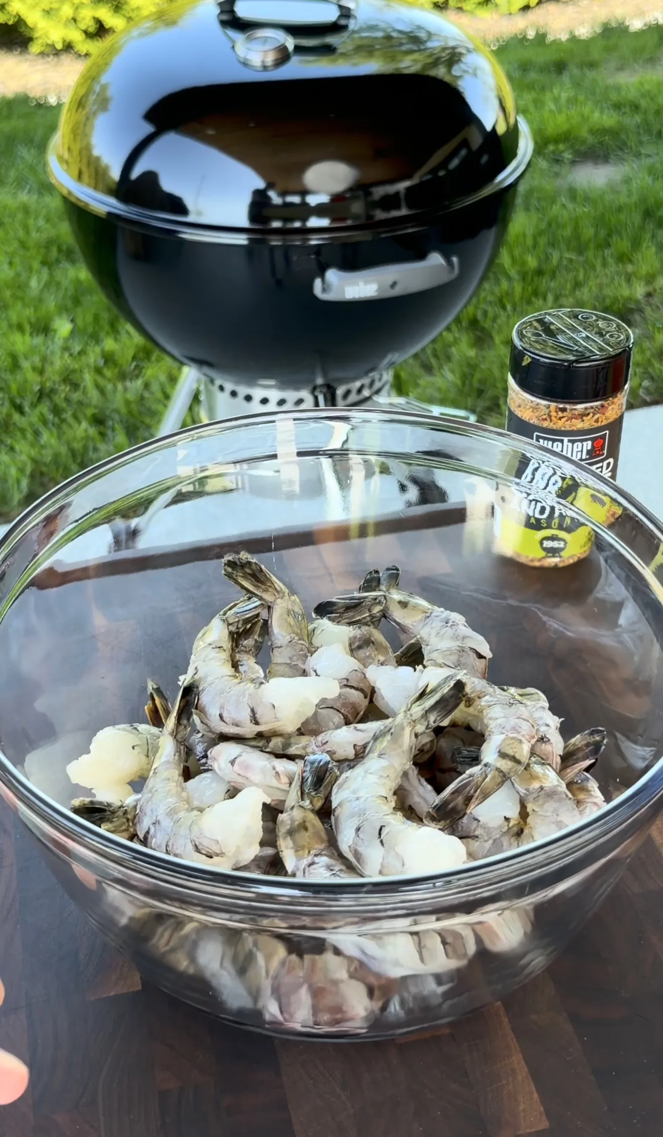 Raw shrimp in glass bowl next to Weber grill with seasoning for grilled shrimp skewers preparation