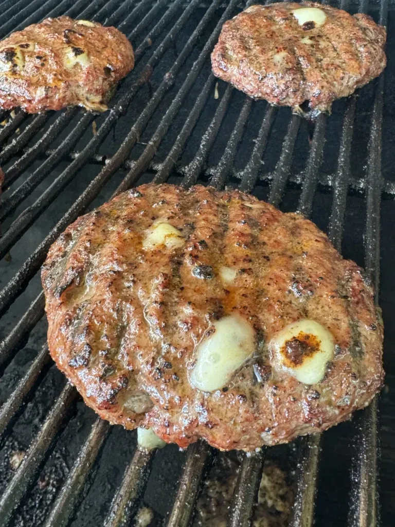 Three seasoned Cajun burger patties cooking on black grill grates with hickory smoke