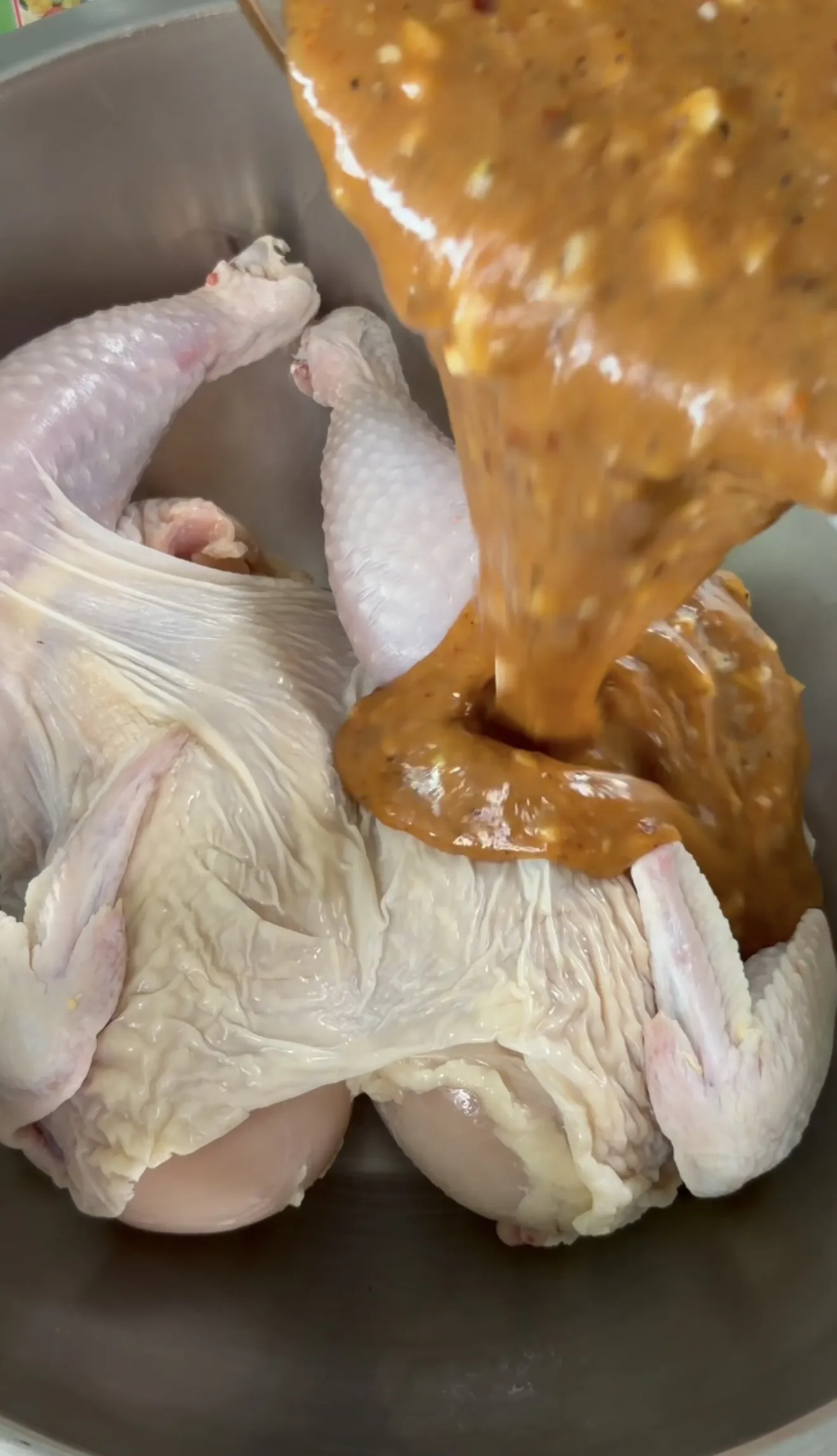 Raw spatchcock chicken being coated with golden marinade sauce in a metal bowl