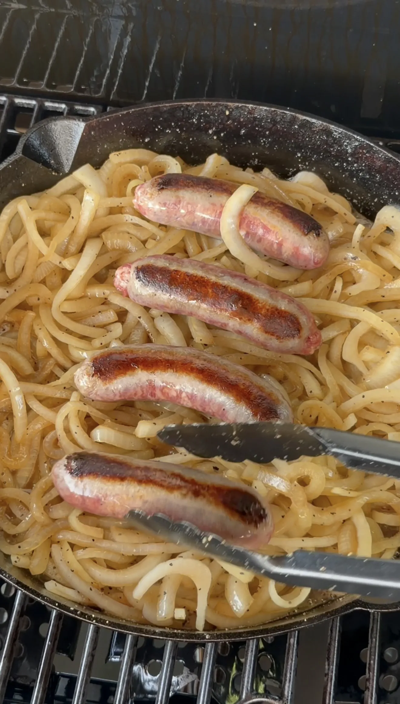 Four beer brats with golden caramelized onions finishing in a cast iron skillet on a grill