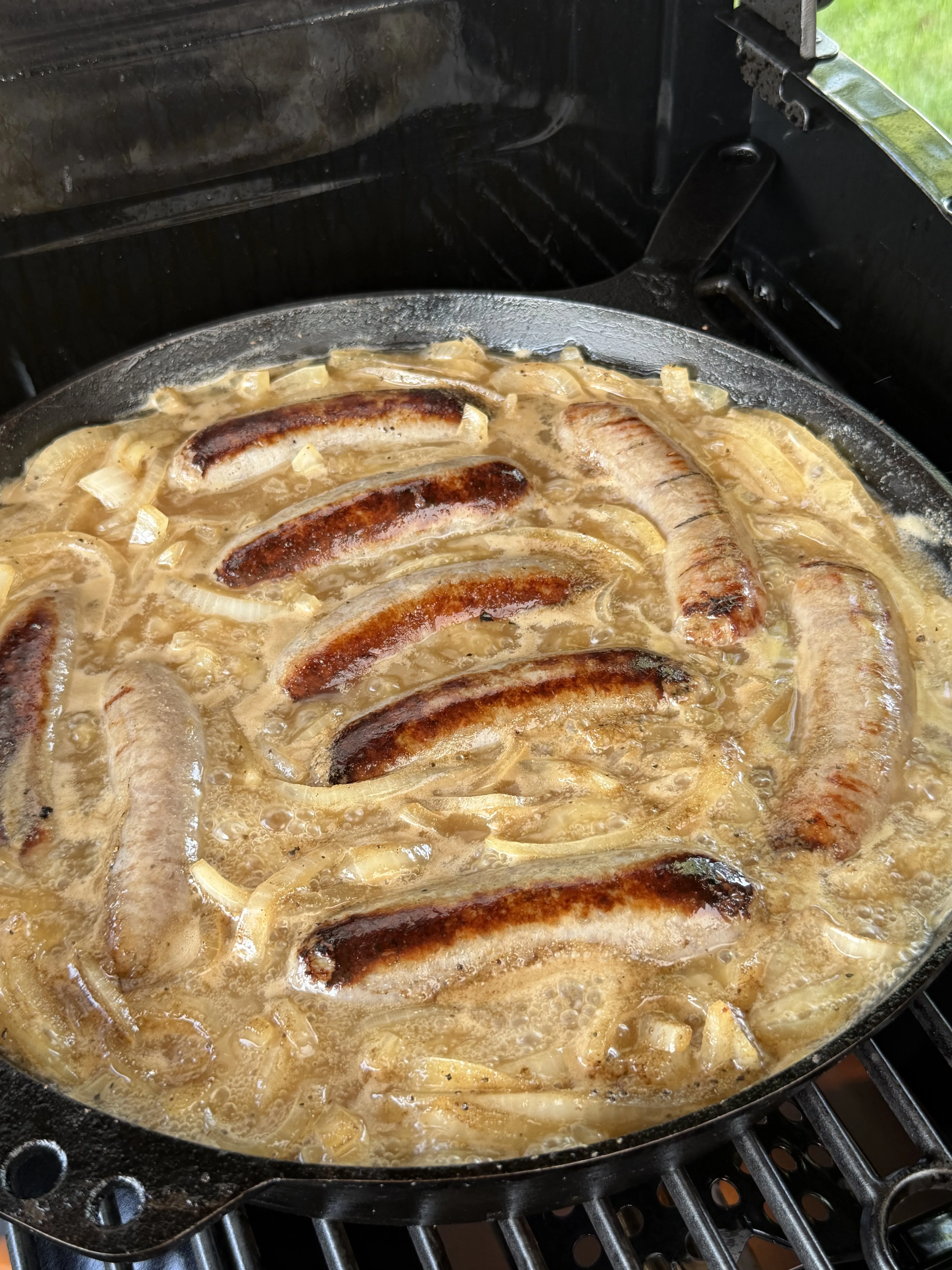 Beer brats simmering with caramelized onions in a cast iron skillet on an outdoor grill