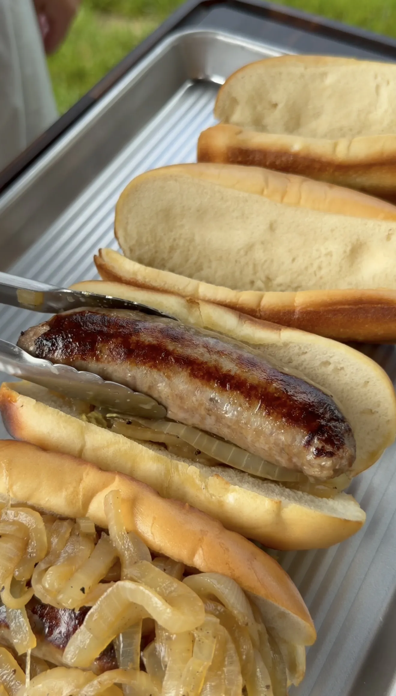 Grilled beer brats being placed into toasted buns with golden caramelized onions